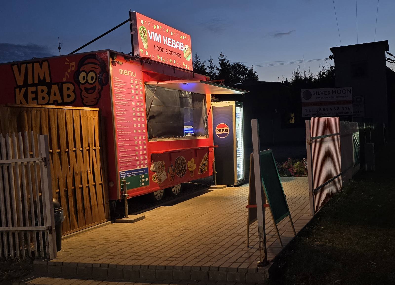 Czerwony food truck VIM Kebab w Łasku, świecący o zmierzchu, z czerwonym podświetlonym menu i lodówką Pepsi.