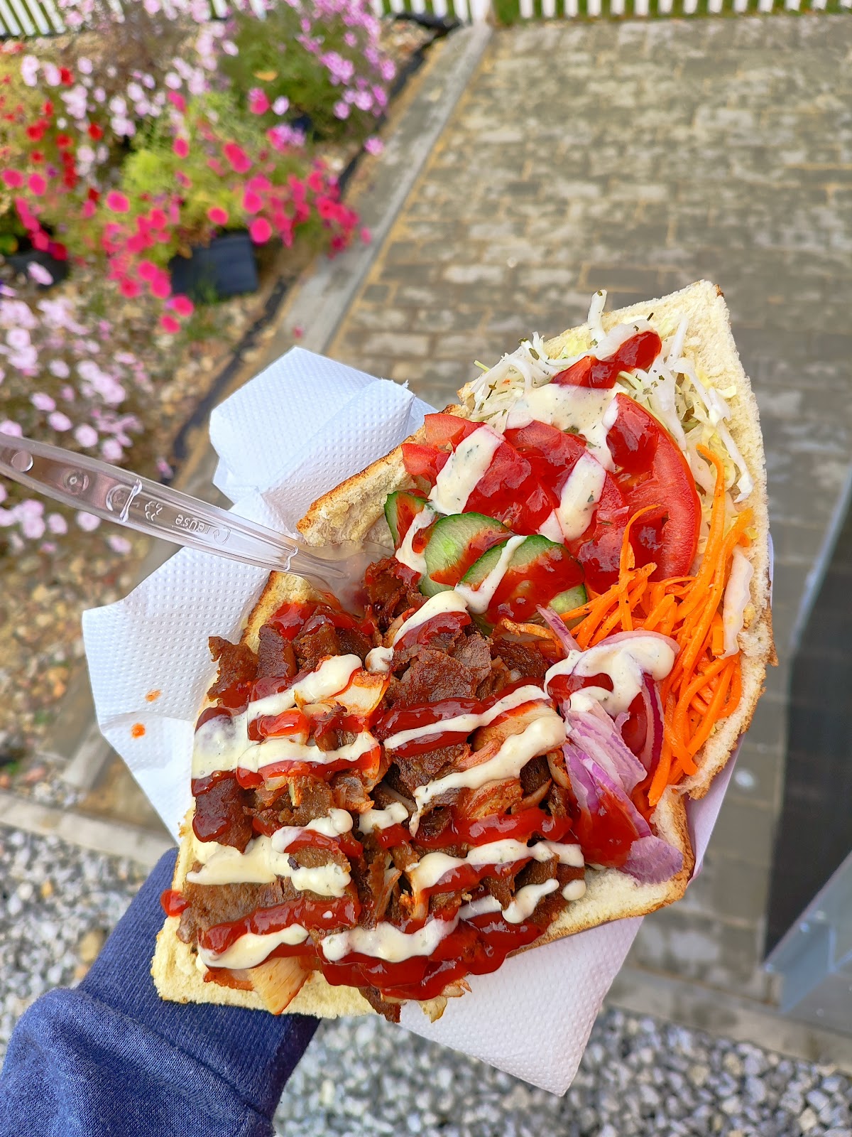 Kebab w bułce VIM Kebab z mięsem, pomidorem, ogórkiem, czerwoną cebulą, marchewką, sosem białym i pikantnym, na tle czerwonych kwiatów.