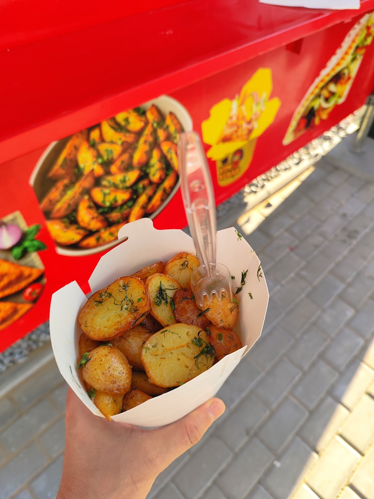 Porcja młodych ziemniaków z koperkiem na tle czerwonego boku food trucka i kolorowych zdjęć dań.
